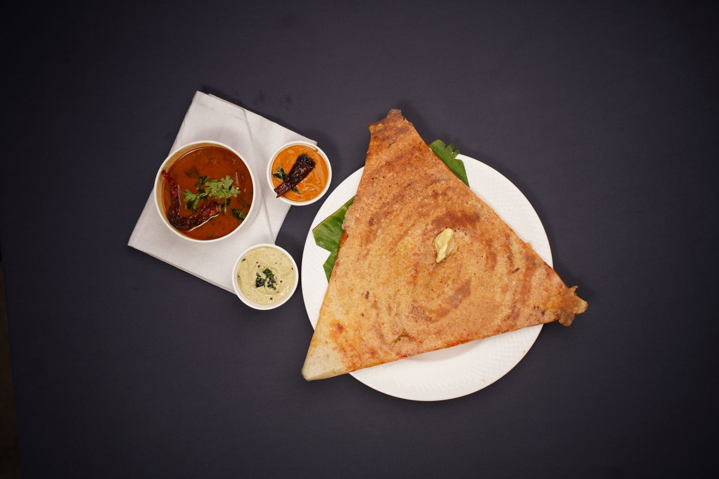 A masala dosa on a steel plate with sambar and two chutneys alongside.