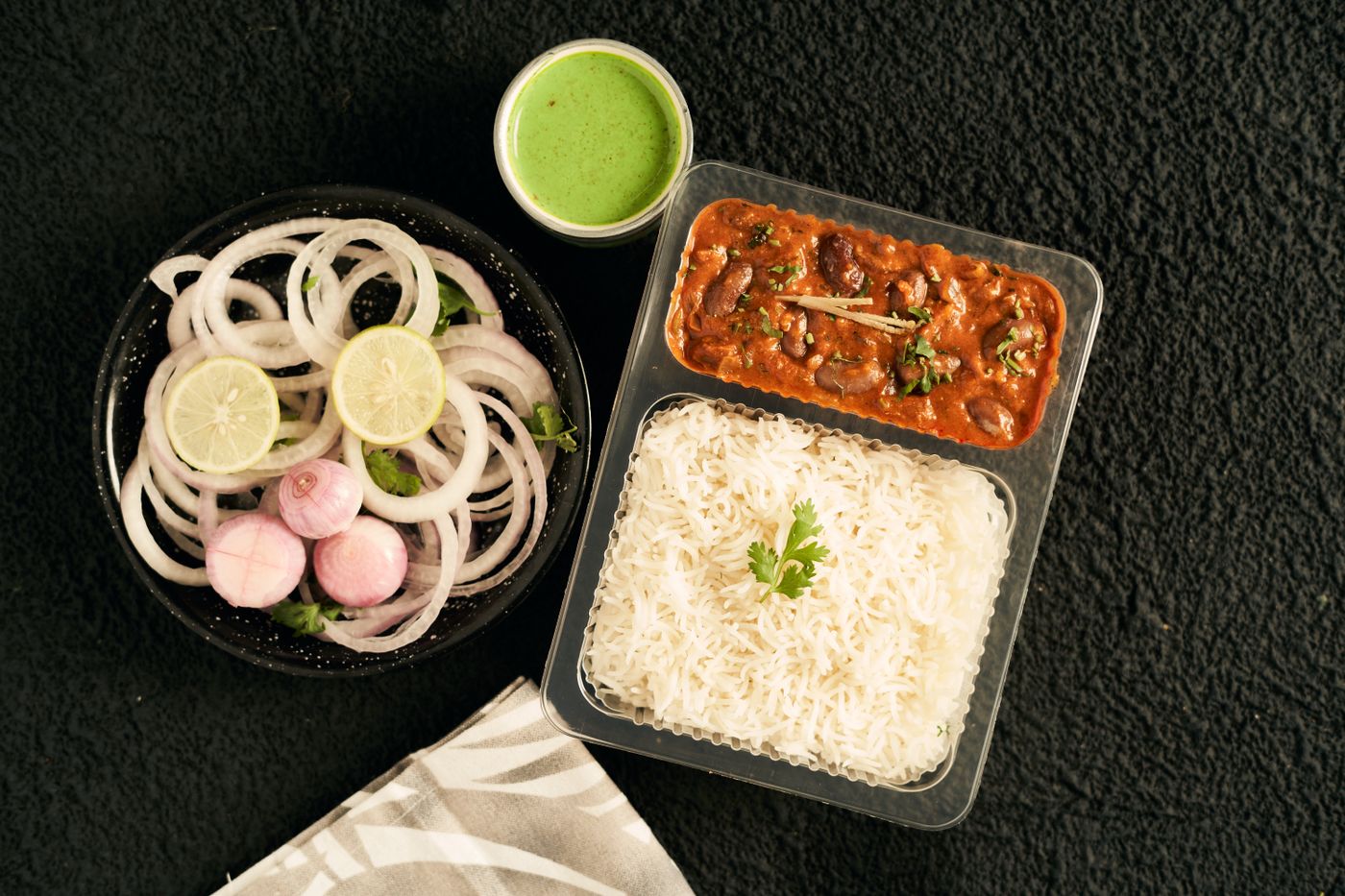 Rajma chawal in a takeaway container, with green chutney and a bowl of sliced
                  onion and lime alongside.