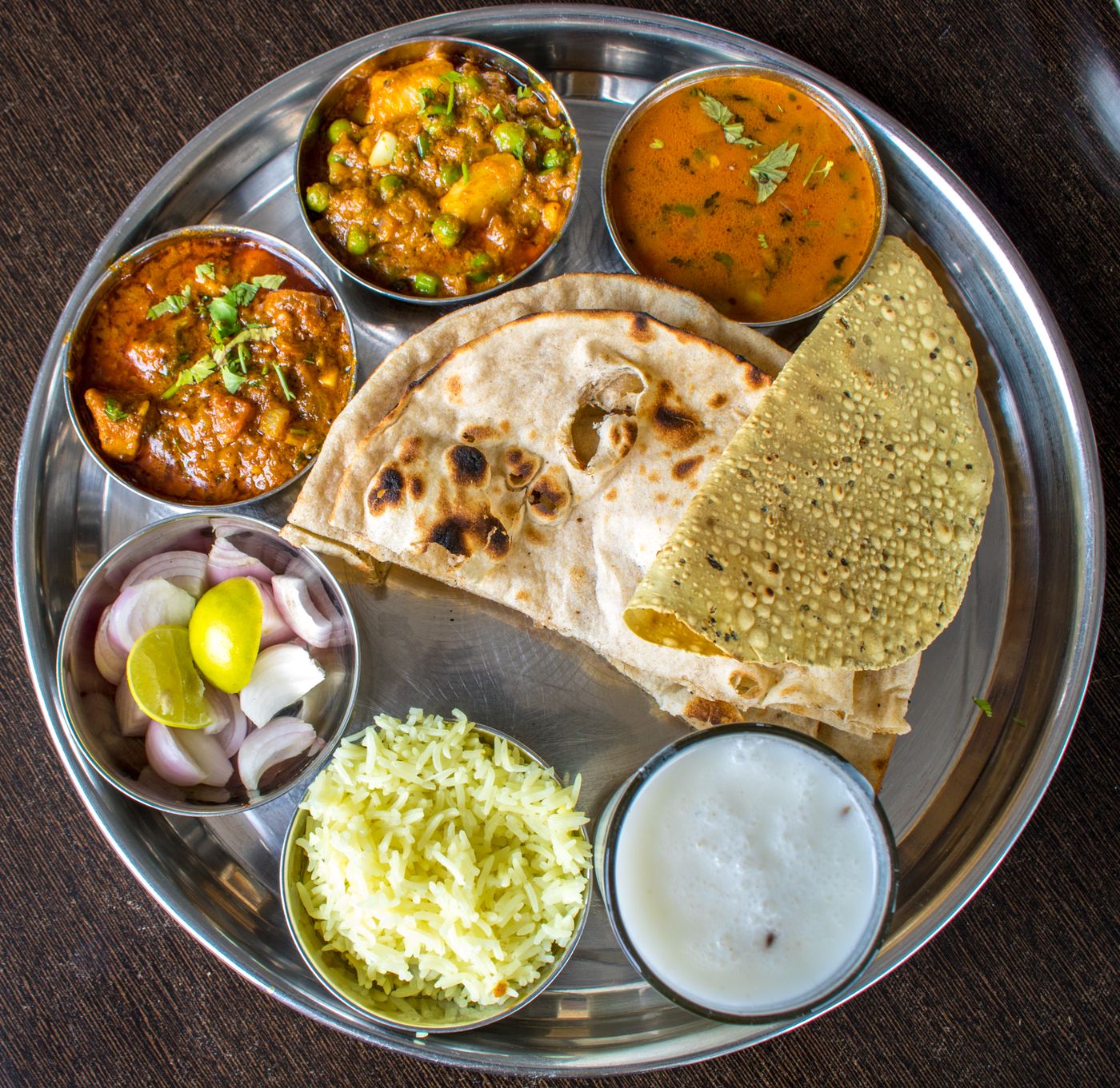 A steel thali holding two vegetable curries, dal, rice, roti, papad, sliced
                      onion and a glass of buttermilk, each in its own katori.